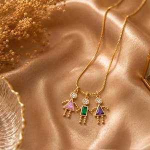 May include: A gold-toned necklace featuring three pendant charms shaped like figures. Each figure has a round clear stone head and a geometric body set with a pink triangular stone, a green rectangular stone, and a purple triangular stone.
