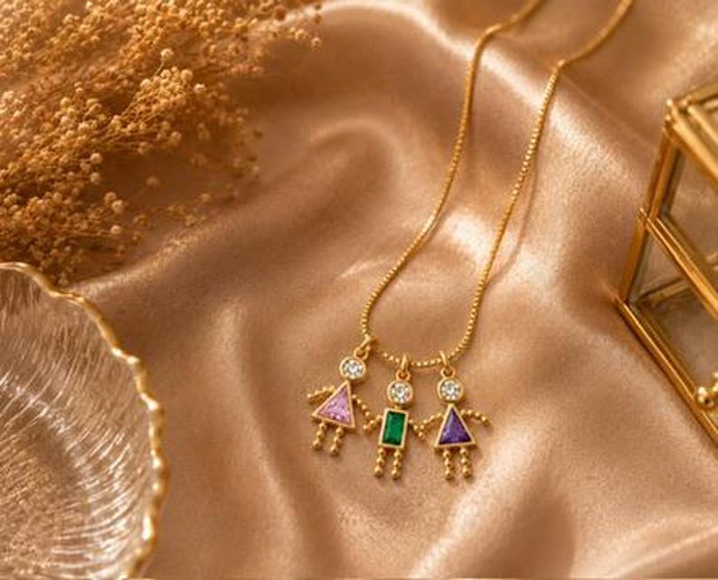 May include: A gold-toned necklace featuring three pendant charms shaped like figures. Each figure has a round clear stone head and a geometric body set with a pink triangular stone, a green rectangular stone, and a purple triangular stone.