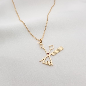May include: Gold-colored necklace with a pendant featuring scissors, a comb, and the name "Lee". The scissors and comb are detailed, and a small red gem is set in the center. The chain is delicate.