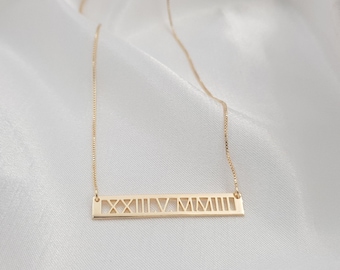 May include: A gold-colored necklace with a rectangular pendant featuring Roman numerals. The pendant is suspended from a delicate chain, resting on a white, textured surface. The Roman numerals are cut out of the pendant.