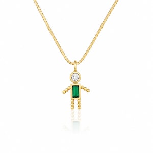 May include: A gold box chain necklace featuring a pendant shaped like a small figure. The pendant has a round clear stone for the head and a rectangular green stone for the torso, with beaded gold arms and legs extending from the central body.