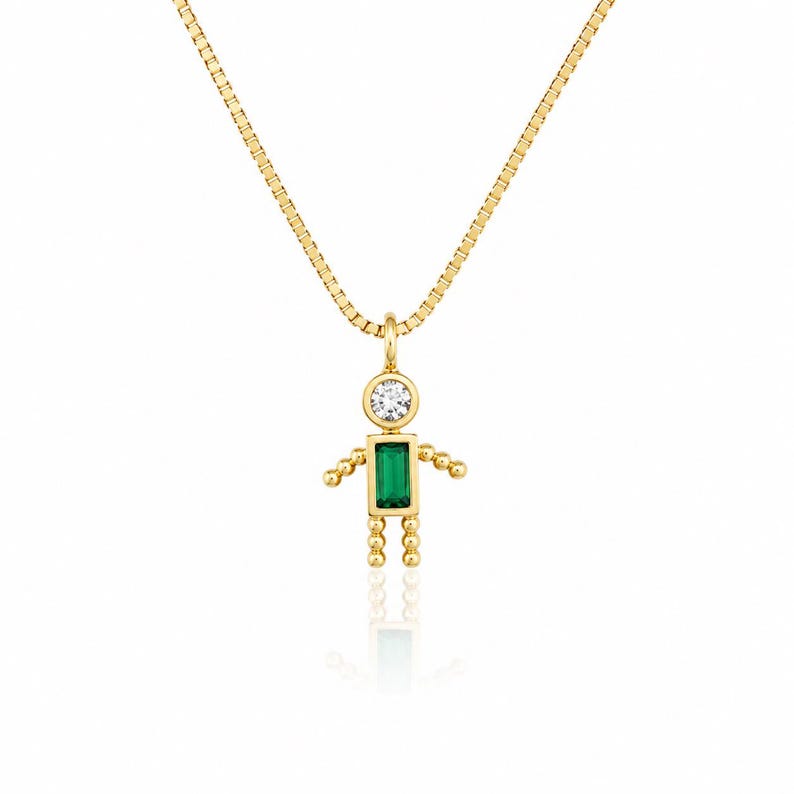 May include: A gold box chain necklace featuring a pendant shaped like a small figure. The pendant has a round clear stone for the head and a rectangular green stone for the torso, with beaded gold arms and legs extending from the central body.