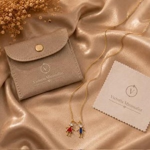 May include: A gold-tone chain necklace featuring two small pendants shaped like a girl in a red dress and a boy in a blue outfit, each with a clear stone accent. The set includes a beige suede-like pouch and a cleaning cloth with the text Victoria Minimalist.
