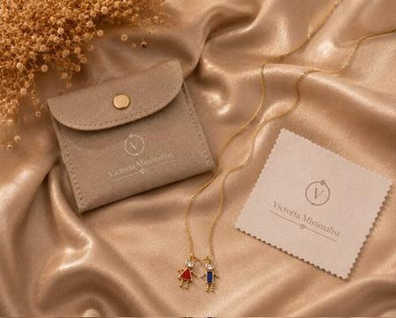 May include: A gold-tone chain necklace featuring two small pendants shaped like a girl in a red dress and a boy in a blue outfit, each with a clear stone accent. The set includes a beige suede-like pouch and a cleaning cloth with the text Victoria Minimalist.