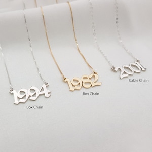 May include: Three necklaces with dates: 1994, 1982, and 2001. The 1994 necklace is silver with a box chain. The 1982 necklace is gold with a box chain. The 2001 necklace is silver with a cable chain. The necklaces are displayed on a white surface.