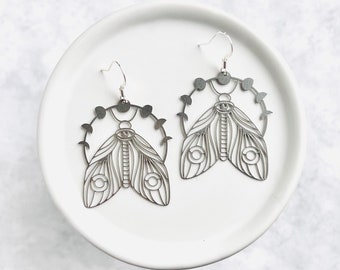 May include: Silver earrings featuring a delicate moth design with a circle of moons surrounding the moth. The moth has intricate wings and a large eye in the centre.