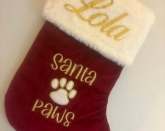 May include: A red Christmas stocking with a white faux fur cuff. The name "Lola" is embroidered in gold script on the cuff. Below, "Santa Paws" and a paw print are embroidered in gold.