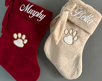 May include: Two holiday stockings, one deep red velvet and one beige faux fur, each featuring a side bow, a white embroidered paw print, and a personalized name in white script. The red stocking reads Murphy and the beige stocking reads Bella.