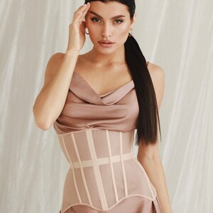 May include: A mauve satin slip dress featuring a cowl neckline and thin straps, paired with a structured beige corset overlay. The corset is designed with vertical boning channels and a curved hemline that sits over the satin fabric of the dress.