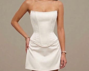 May include: A strapless, off-white mini dress featuring a structured corset-style bodice with vertical seams and a defined waistline. The garment has a smooth, satin-like finish and a straight-cut skirt that falls above the knee, creating a clean silhouette.