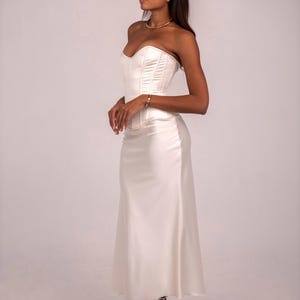 May include: A cream-colored satin corset top paired with a matching floor-length satin skirt. The corset features structured boning and a sweetheart neckline, while the skirt has a smooth, flowing silhouette that falls to the ankles.