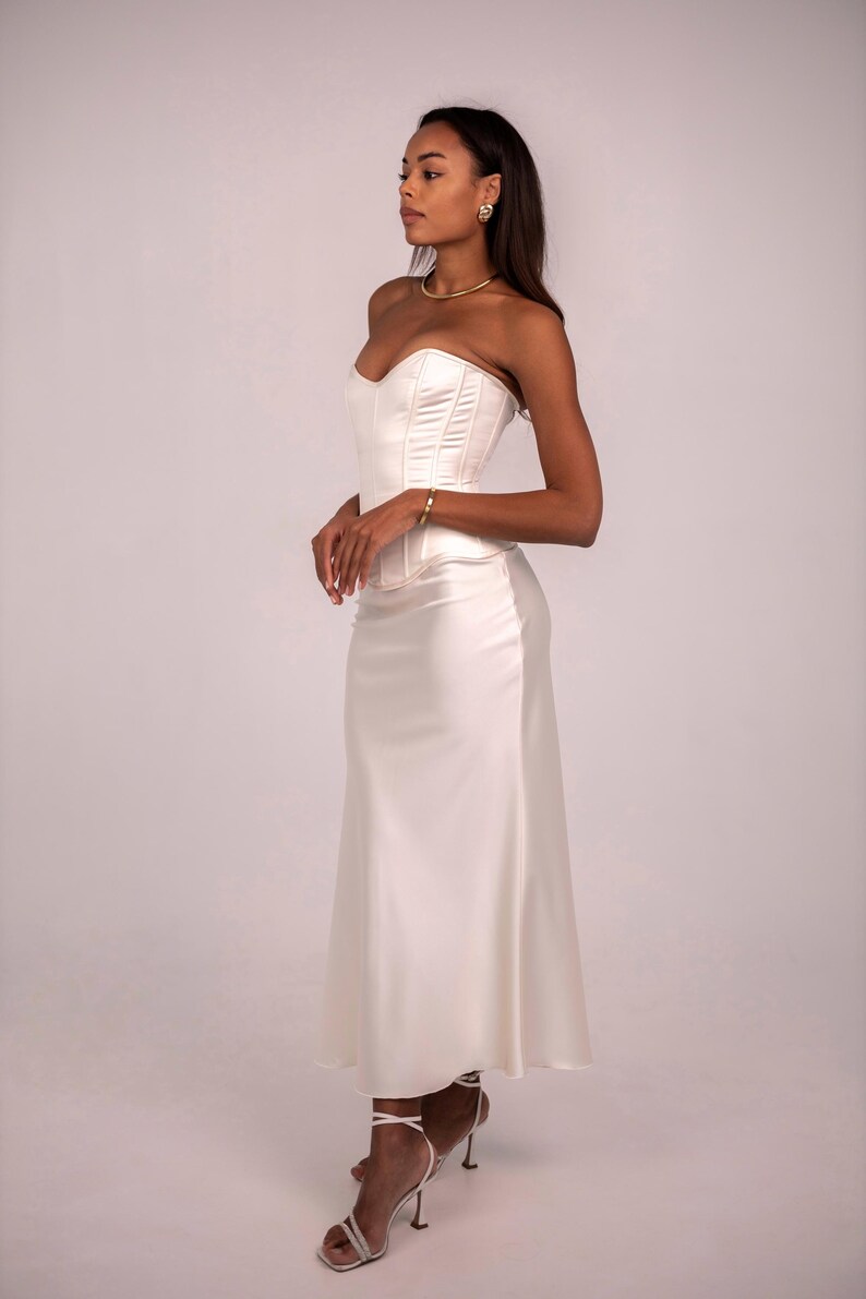 May include: A cream-colored satin corset top paired with a matching floor-length satin skirt. The corset features structured boning and a sweetheart neckline, while the skirt has a smooth, flowing silhouette that falls to the ankles.