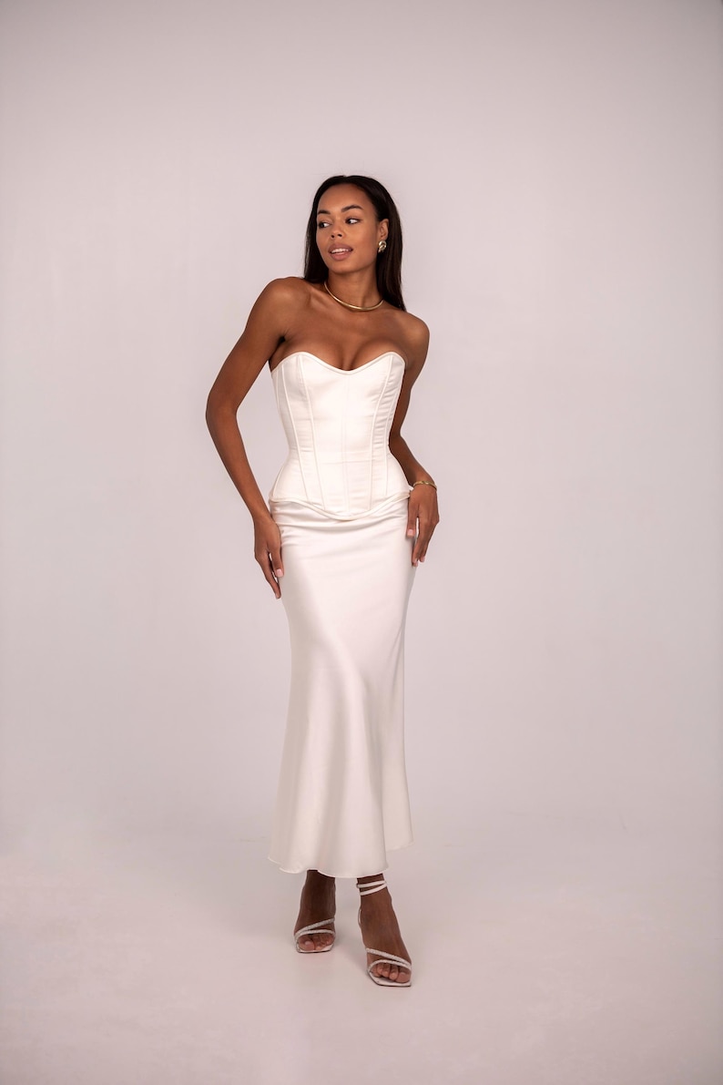 May include: A strapless ivory corset top with vertical boning seams paired with a matching floor-length satin skirt. The ensemble features a sweetheart neckline on the bodice and a smooth, flowing silhouette that extends to the ankles.