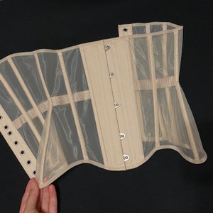 May include: A beige mesh corset with metal eyelets and a busk closure. The corset is made of a sheer fabric and has a scalloped edge.
