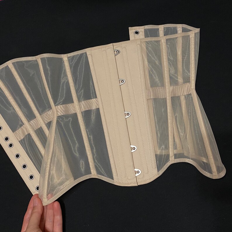 May include: A beige mesh corset with metal eyelets and a busk closure. The corset is made of a sheer fabric and has a scalloped edge.