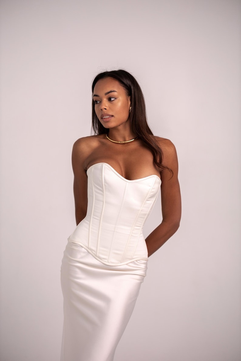 May include: A strapless ivory corset top featuring vertical boning channels and a sweetheart neckline, paired with a matching ivory satin skirt. The garment has a structured bodice with a curved hemline that sits at the waist, creating a sleek silhouette.
