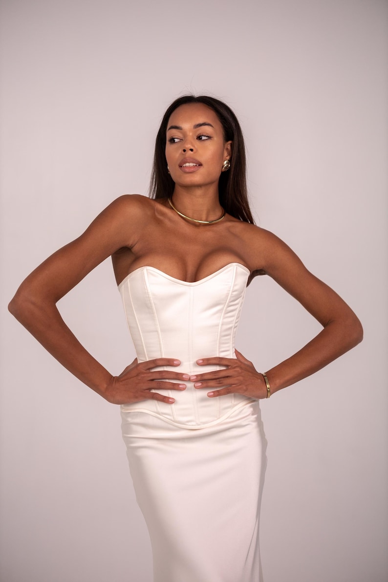 May include: A strapless, ivory-colored corset top featuring vertical seam detailing and a sweetheart neckline. The garment is constructed from a smooth, satin-like fabric and is paired with a matching floor-length skirt in the same ivory tone.