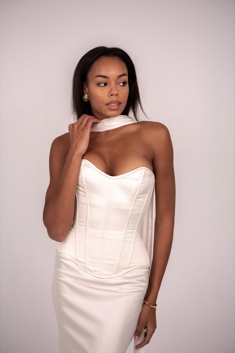 May include: A white strapless corset top featuring structured vertical boning and a sweetheart neckline, paired with a matching white scarf draped around the neck. The garment is crafted from a smooth, satin-like fabric with a clean, minimalist finish.