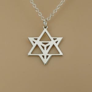 Può includere: Una collana color argento con un ciondolo a forma di Stella di David. Il ciondolo è una stella a sei punte con un design geometrico. La catena è un modello delicato e a maglie. Lo sfondo è di colore neutro.
