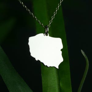 May include: A silver-colored pendant necklace featuring a polished outline of Poland. The pendant hangs from a delicate chain, set against a dark green background. The necklace is a simple, elegant piece of jewelry.