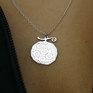 May include: A silver pendant necklace with a fruit-shaped charm, decorated with a swirling pattern. The necklace hangs from a fine chain. The pendant is shown against a dark background.