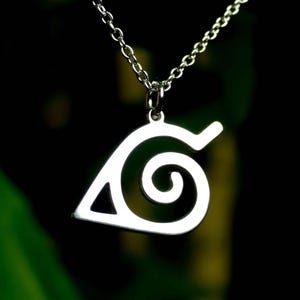 May include: A silver pendant necklace with a leaf design. The pendant is a stylized leaf shape with a spiral and a curved top. The chain is delicate, and the background is dark green and black.