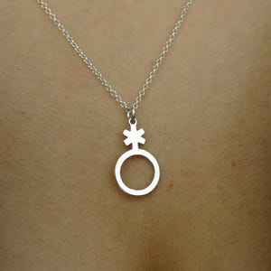 Könnte beinhalten: Eine silberfarbene Halskette mit einem Anhänger, der ein genderqueeres Symbol zeigt. Der Anhänger ist ein Kreis mit einem kreuzartigen Sternchen darüber. Die Halskette wird vor einem dunklen Hintergrund gezeigt.