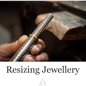 Könnte beinhalten: Ein goldener Ring wird auf einem Metall-Ringmaß gemessen. Der Ringmaß wird von einer Hand gehalten. Das Bild ist eine Nahaufnahme des Rings und des Ringmaßes.