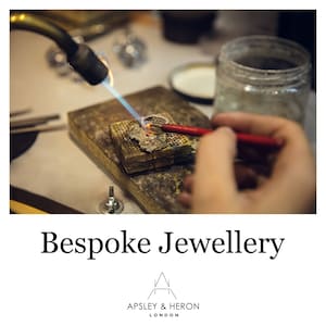 Puede incluir: Primer plano de un joyero que utiliza un soplete para calentar un anillo de plata en un banco de trabajo de metal. El anillo se mantiene en su lugar con una herramienta roja. El texto "Bespoke Jewellery" y "Apsley & Heron London" es visible debajo de la imagen.