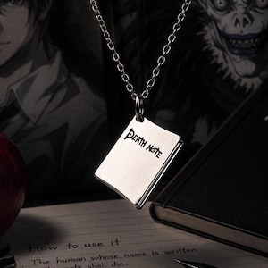 May include: A silver pendant necklace featuring a small, rectangular charm engraved with "DEATH NOTE". The charm hangs from a delicate chain. A red apple and a black notebook with the words "DEATH NOTE" are also visible, along with handwritten text.