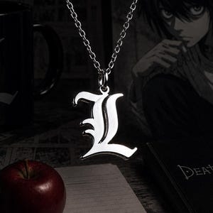 Pode incluir: Um colar com pingente prateado com a letra "L" estilizada. A corrente é fina e delicada. Uma caneca preta com o logotipo "L" branco, uma maçã vermelha e um livro "Death Note" também são visíveis.