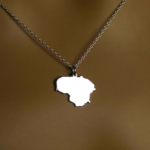 May include: A silver-colored necklace featuring a pendant in the shape of the country of Lithuania. The pendant is suspended from a delicate chain. The necklace is displayed against a brown background.
