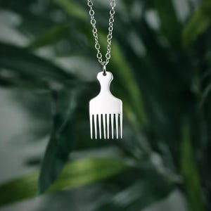 May include: A silver-colored pendant necklace featuring a comb-shaped charm. The charm has a handle and multiple tines, suspended from a delicate chain. The background is a blurred view of green foliage.
