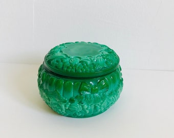 May include: A round, emerald green trinket box with a matching lid. The box and lid are adorned with a detailed floral and leaf pattern. The box is sitting on a white surface.