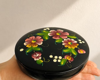 Può includere: Una scatola rotonda nera con coperchio caratterizzata da un motivo floreale dipinto a mano sulla parte superiore. Il design include fiori rosa e gialli con foglie verdi e piccoli punti bianchi disposti in un motivo circolare sulla superficie scura.
