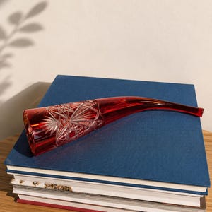 May include: A ruby red glass horn-shaped vase featuring intricate starburst and geometric cut patterns on the wider end. The object is elongated and curved, tapering to a narrow opening at the tip, showcasing a polished, translucent finish.