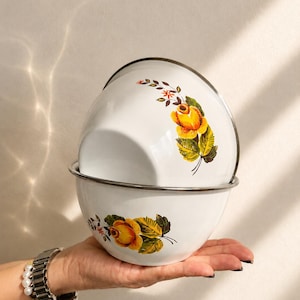 May include: Two white enamel bowls with a silver rim, each decorated with a floral pattern featuring yellow roses and green leaves. The bowls are stacked, with one resting inside the other, showcasing the smooth, glossy finish of the metal surface.