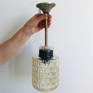 May include: A pendant light fixture featuring a cylindrical, amber-tinted glass shade with a textured, repeating teardrop pattern. The shade is attached to a brass-colored metal rod and a fluted, silver-toned ceiling canopy mount.