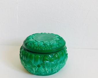 May include: A round, emerald green glass jar with a matching lid. The exterior features a detailed, raised relief pattern of clusters of grapes, leaves, and decorative bows encircling the body and the top surface of the lid.
