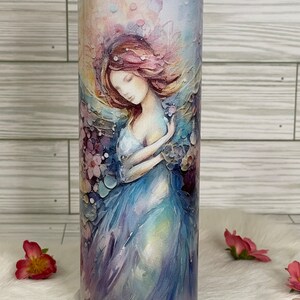 May include: A stainless steel tumbler with a watercolor painting of a woman in a flowing blue dress. The artwork features soft pink, purple, and blue hues, with a dreamy, ethereal quality. The tumbler is designed for cold beverages.
