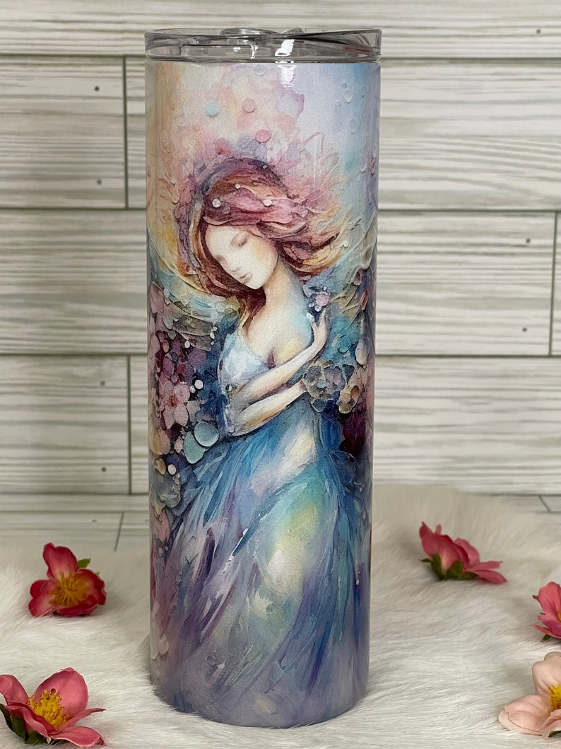 May include: A stainless steel tumbler with a watercolor painting of a woman in a flowing blue dress. The artwork features soft pink, purple, and blue hues, with a dreamy, ethereal quality. The tumbler is designed for cold beverages.