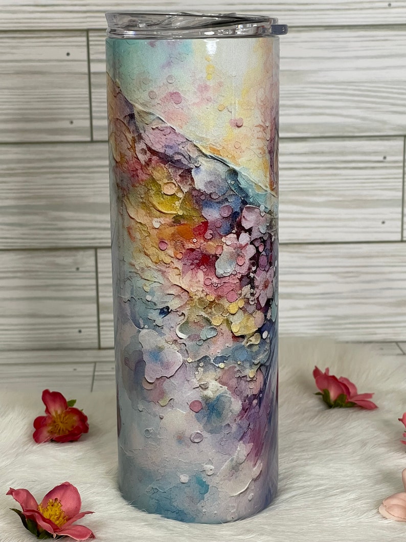 May include: A tall, insulated tumbler with a colorful, abstract watercolor design. The design features a blend of pastel colors, including pink, blue, yellow, and purple, creating a textured, artistic appearance. The tumbler has a clear lid and is suitable for cold beverages.