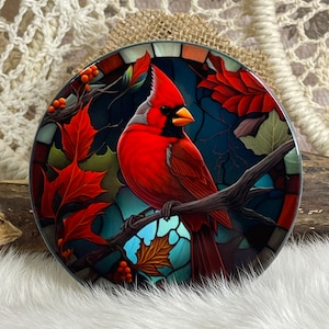 May include: A round decorative coaster featuring a vibrant red cardinal perched on a branch, surrounded by autumn leaves. The coaster has a stained-glass effect with a dark blue background and a glossy finish. The design includes orange berries and a detailed illustration.