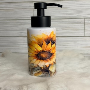 May include: A cylindrical soap dispenser with a black pump top. The dispenser is decorated with a watercolor-style sunflower design in shades of yellow, orange, and brown. The background is white.