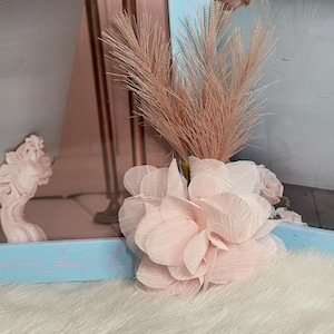 May include: A pink fabric flower with a light brown dried grass bouquet. The flower is on a white fluffy surface.