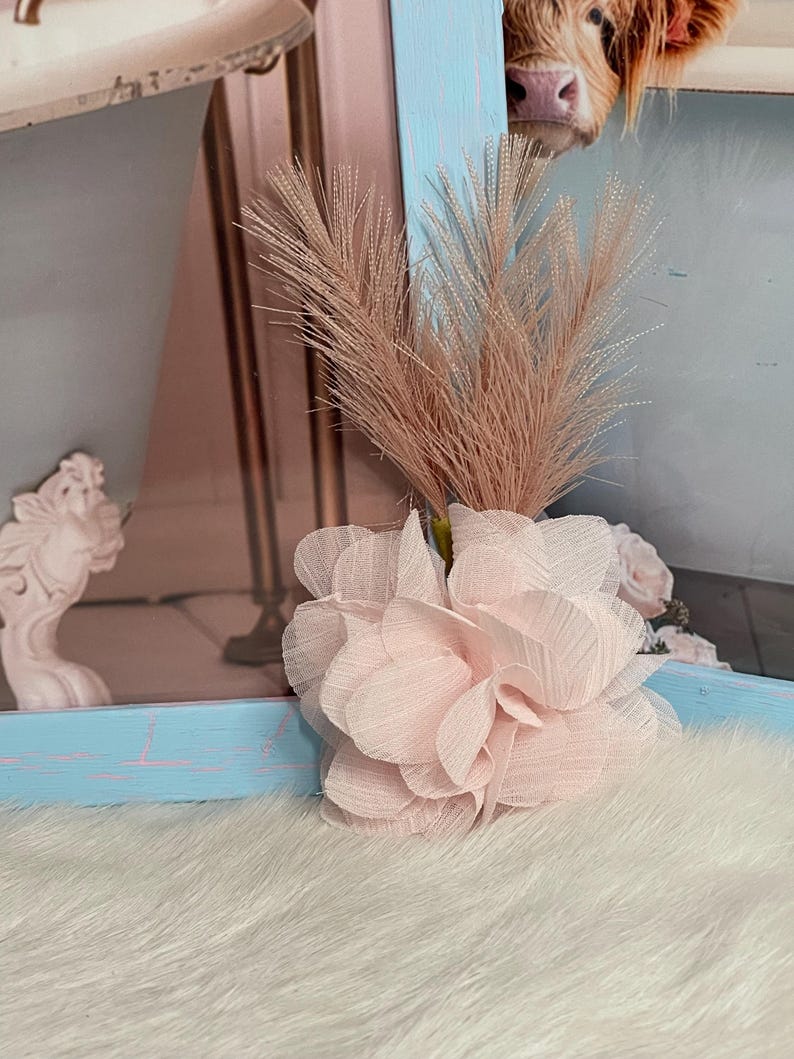 May include: A pink fabric flower with a light brown dried grass bouquet. The flower is on a white fluffy surface.