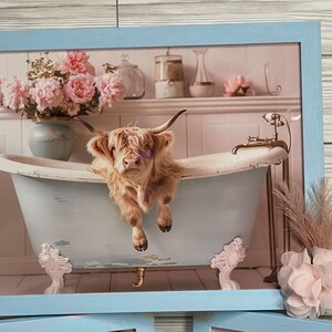May include: A Highland cow relaxes in a vintage clawfoot bathtub with pink flowers in the background. The cow is brown and white and has a fluffy coat. The bathtub is blue and white and has gold accents. The flowers are in a vase on a shelf above the bathtub.