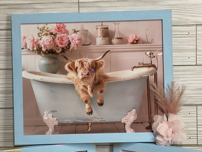 May include: A Highland cow relaxes in a vintage clawfoot bathtub with pink flowers in the background. The cow is brown and white and has a fluffy coat. The bathtub is blue and white and has gold accents. The flowers are in a vase on a shelf above the bathtub.