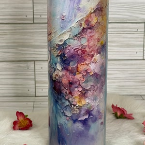 May include: A tall, cylindrical tumbler with a clear lid. The tumbler is decorated with an abstract watercolor design in shades of purple, pink, blue, and yellow. The design has a textured appearance, with a mix of soft and defined shapes.