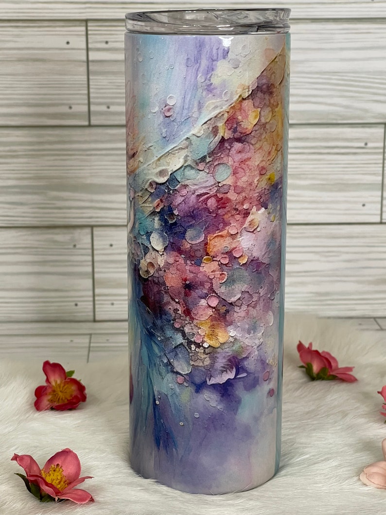 May include: A tall, cylindrical tumbler with a clear lid. The tumbler is decorated with an abstract watercolor design in shades of purple, pink, blue, and yellow. The design has a textured appearance, with a mix of soft and defined shapes.